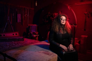 Sunny Megatron Kink - Curtis Joe Walker, Photo Bang Bang 2021 (Sunny sitting in a dark dungeon space with impact play equipment hanging behind her on the walls